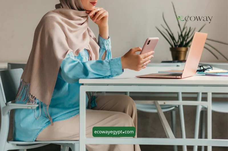 امرأة ترتدي الحجاب تجلس إلى مكتب يضم جهاز كمبيوتر محمولاً وهاتفاً. ترتدي قميصاً أزرق وسروالاً بلون بيج. النص: ecoway, ecoway egypt.com.